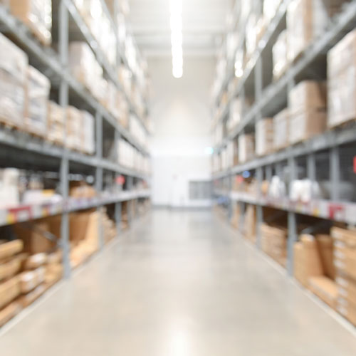 Warehouse Supply Industry Market Blurry perspective of a large warehouse aisle with shelves stacked with boxes and goods.
