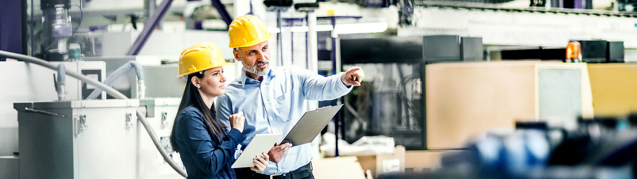 Two project managers standing in modern industrial factory, looking document in clipboard. Manufacturing facility with robotics, robotic arms and automation